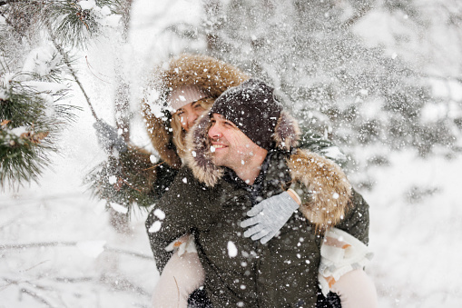 Cheerful young couple having fun in winter forest while piggybacking and shaking branch making snow fall