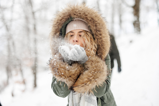 Cheerful young woman blowing snow from her hands in winter forest