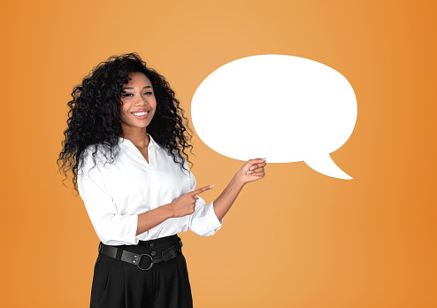 African American businesswoman wearing formal wear is standing pointing up with fingers to speech bubble icon near empty orange wall in background. Concept of model, successful business person, show