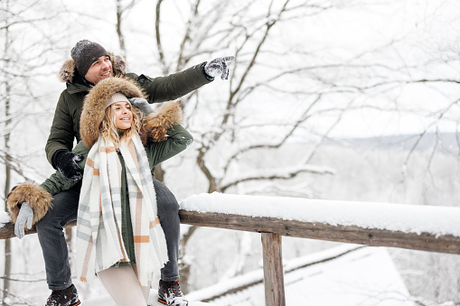 Cheerful young couple standing on wooden terrace and enjoying the view on forest on cold winter day