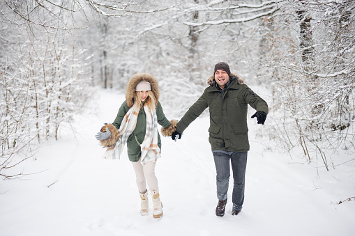 Cheerful couple holding hands and happily running in snowy forest on cold winter day