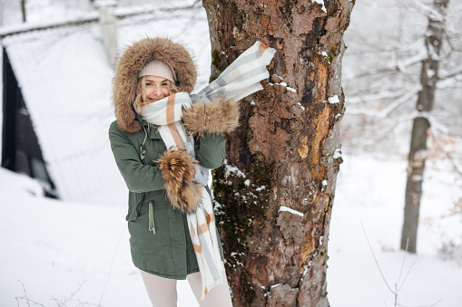 Beautiful young woman in hooded jacket standing in snowy forest on cold winter day