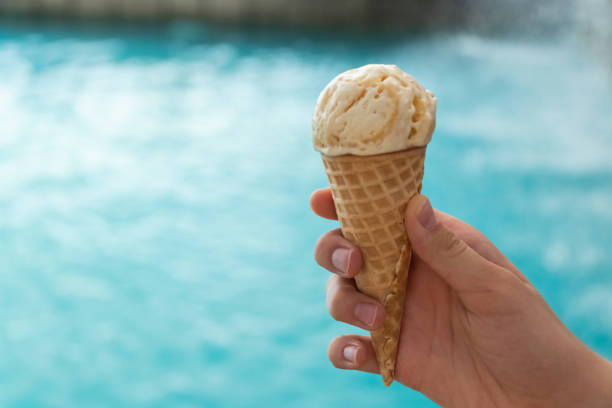 young hand holds an ice cream cone on the background of the blue water of the sea. the concept of relaxation by the sea on a bright, hot and sunny day stock photo