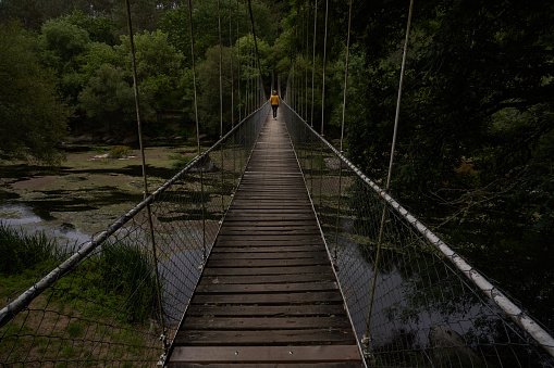 Woman silhouette in the distance on a wooden suspension bridge with metal fence over the Miño river in Spain surrounded by nature.