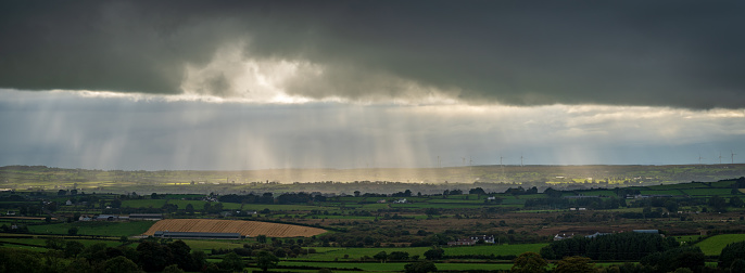 Panoramic wide view of moody dark rain clouds with sunbeams and rays of light shining through illuminating the countryside near Dunloy, County Antrim, Northern Ireland. With rolling hills in foreground and a variety of agricultural fields, trees, hedges and buildings.