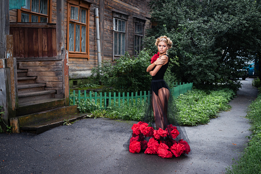 Beautiful young woman in black dress with red flowers is standing at the backyard of old rotten wooden building under the rain