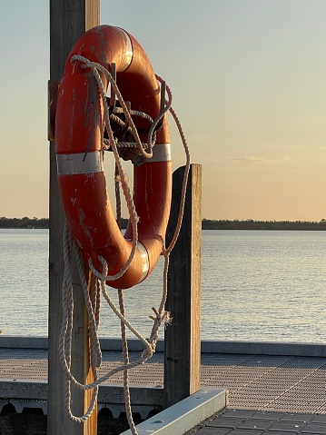 Red lifebuoy ring with ropes hooked as a safety ring at the boat deck in Loch sport Lake with a blue yellow orange sunset and land in the background