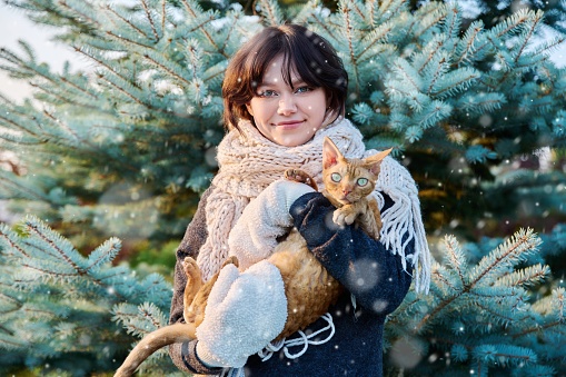 Young happy female in snowy weather cat in arms looking at camera, outdoor near Christmas tree. Teenage female in knitted hat scarf mittens with pet snowflakes. Season winter, beauty New Year holidays