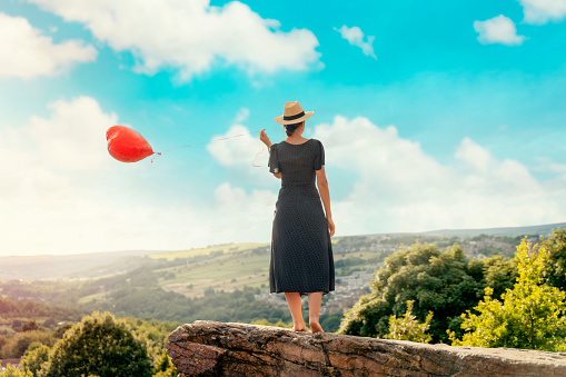 woman holding a red balloon in the shape of a heart on top of the hill against the blue cloudy sky