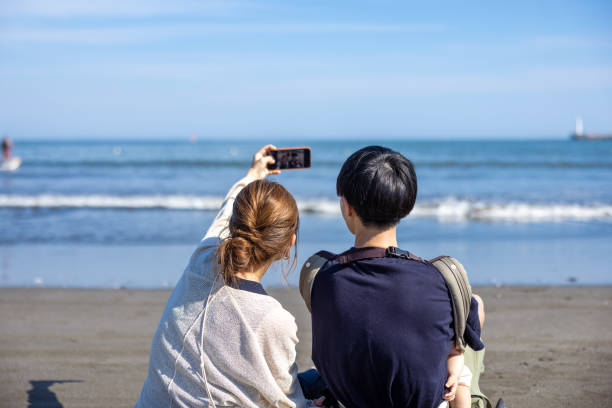 ビーチに座って自撮り写真を撮っている家族の背面図 - 夫婦 ストックフォトと画像