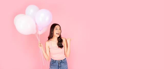 Cheerful woman holding balloons point up to copy space in excitement Expressive facial expressions Beautiful girl enjoy holiday Celebrate for party or special day Use for advertising Pink background