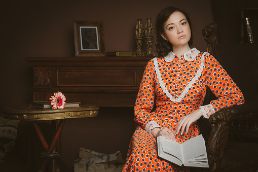 Retro-styled portrait of young attractive woman sitting on a chair in old fashioned interior with a book on her knees