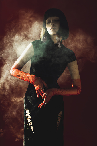 Fabulous brunette woman posing with smoke in studio on red background. Fashion model with pale skin and brown hair in black dress. Gloves on hands. Fashion style.