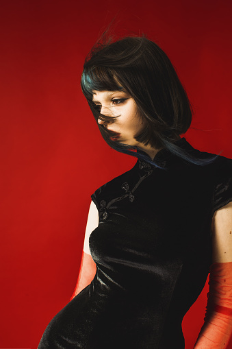 Fabulous brunette woman posing in studio on red background. Fashion model with pale skin and brown hair in black dress. Gloves on hands. Fashion style.