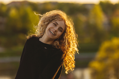 Happy blonde curly beautiful girl in sweater against sunset sky. Charming smile. Close up portrait. Autumn break. Woman enjoying vacation. Girl in nature. Feel happiness. Warm light