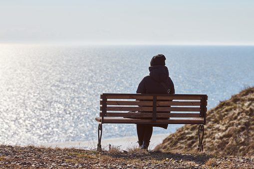 Single girl in a black jacket and hat sitting on bench at cliff at front of sea, peaceful and quiet place for thinking alone, loneliness and loss of loved one concept. Pacifying view of marine horizon
