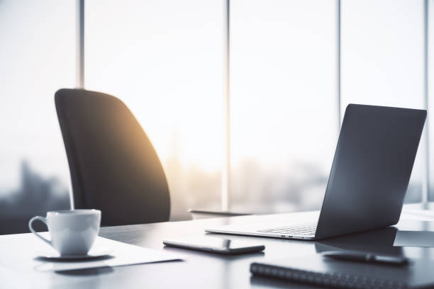 istock Perspective view on sunlit office workspace with modern laptop, tea cup and smartphone on dark table surface and black chair on big window background. 3D rendering 1429690412