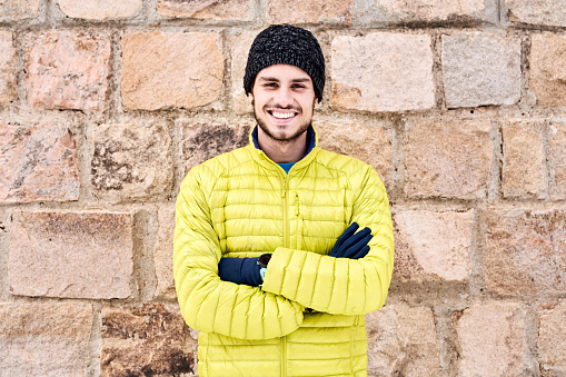 Portrait of good-looking smiling man with beard wearing yellow winter jacket, grey hat and gloves, standing over stone wall with folded arms.
