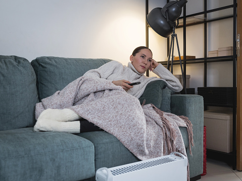 middle aged woman resting  on the couch covered with a blanket and warming herself in cold weather using electric heater