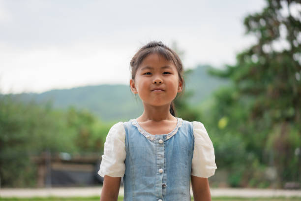 Asian girl observing a small object in hand while standing in a national park, wearing a pink dress and white headband. Child deeply focused on exploring nature and enjoying her surroundings.