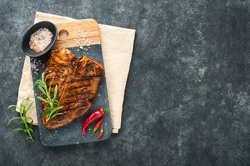 Raw steak t-bone or porterhouse. Steak t-bone or porterhouse in sauce with spices, salt and rosemary on a black ceramic plate on concrete background. Top view. Place for text.