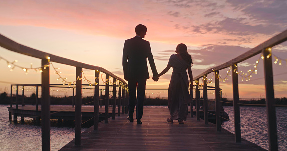 Silhouette couple holding hand at lake or river during summer sunset date outdoor. Man and woman walk on a bridge together with calm water while enjoying romance, love and relation while on vacation