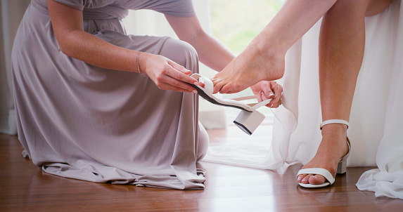 Wedding shoes, helping bride and women prepare for marriage ceremony. Bridesmaid putting heels on woman friends feet for happy event. Bridal party for engagement celebration of people in commitment.