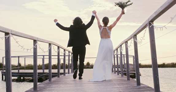 Happy couple celebration after wedding on a bridge at a lake or river together from the back. Bride and groom excited, happiness and married wearing suit and dress during summer with sky background