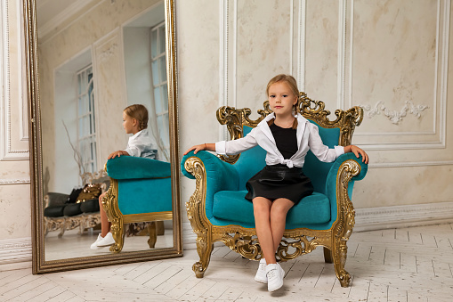 Little princess smiling 4-5 years old blonde girl model in stylish clothe in living room with reflection at mirror, looking at camera. Fashionable young lady model in room, sitting on armchair