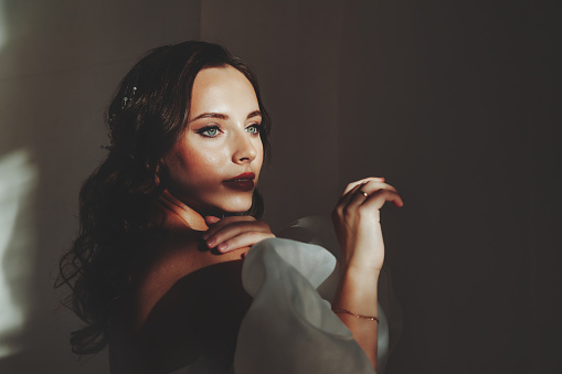 Portrait stylish bride young woman in white wedding dress standing in shadow, looking away. Bride lady in vintage room, grey wall background in studio. Fashion style concept. Copy text space