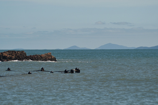 The poles of a netted ocean bath are just visible in a high tide. Rocks are behind the pool, and hills can be seen across the bay. The sky is blue, with faint clouds.