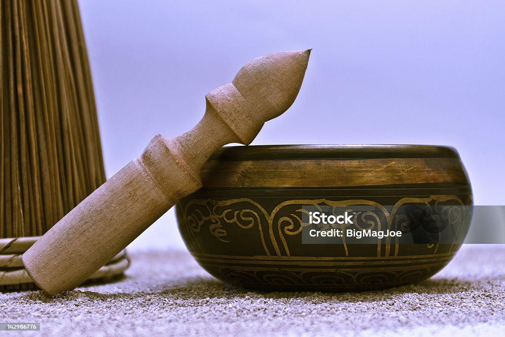 Singing bowl in sand Studio close up of a singing bowl with a wooden stick. Coarse sand on the floor and an wood tree on the side. Gray-blue background. Alternative Therapy Stock Photo