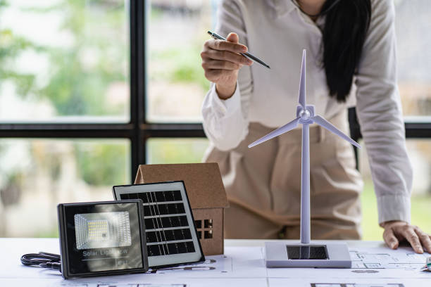 istock Young Asian architect with blueprints working on lapotp in office. Career working hard on wooden table, painting with windmills, designing model of renewable wind house building. and sunlight 1429872705
