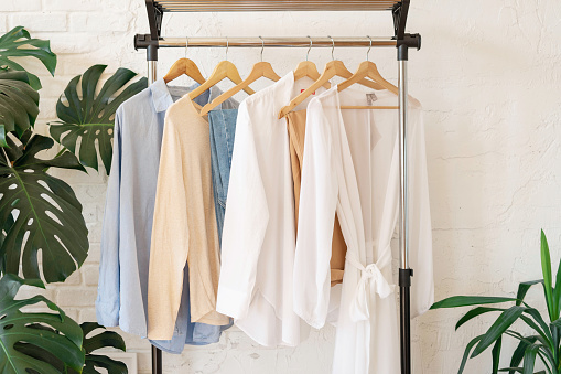 Trendy capsule wardrobe in beige and light blue on a rail rack against the background of a white textured wall with monstera leaves.