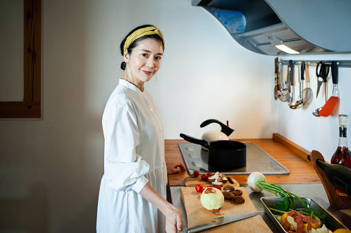 Health and Sustainability.\nA woman wearing a white dress.\nA woman is cutting vegetables.\nNatural modern style kitchen.