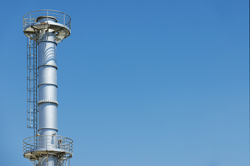 High section of industrial metallic chimney with security railing and ladder, against clear blue sky