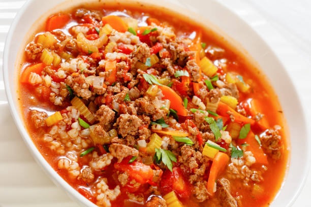Easy hot and hearty Hamburger Soup with barley and vegetables in white bowl on white wood table, horizontal view from above