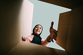 Smiling young Asian Muslim woman opening carboard box while looking at camera. - Presents, delivery and holidays concept