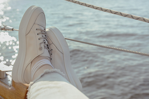 female crossed legs in white sneakers on the deck of yacht at sea, first-person view on sunset, selective focus, copy space