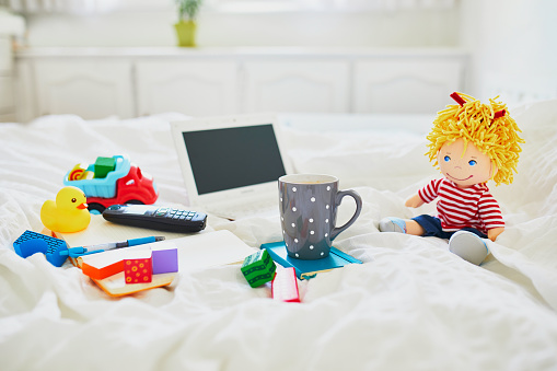Laptop, cup of coffee, notebook, phone and different toys in bed on clean white linens. Freelance, distance learning or work from home with kids concept