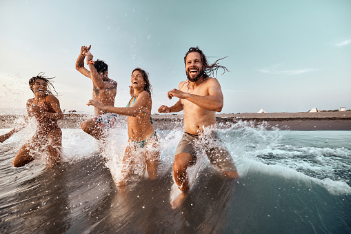 Group of cheerful friends having fun while running through sea in summer day. Copy space.