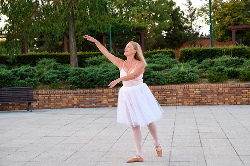 Mature smiling woman dancing ballet in a park at street.