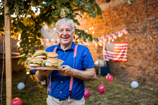 A senior man holding a cutting board with burgers on it at an American national holiday barbecue party