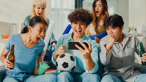 Group of happy Asian teenage people sit on couch watch cheering sport soccer games together on smartphone. Shouting strike goal excited scream, facial expression concept.