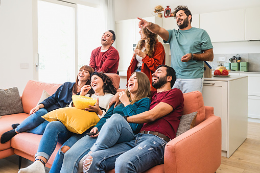A group of young adults people enjoy watching funny content such as comedy movie on TV while eating popcorn, the bearded guy points on the television screen, while friends laugh out loud