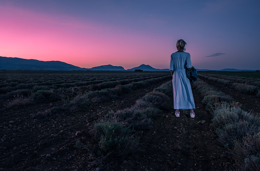 Mature woman in a blue dress looking towards sunrise over La Grande Marge at the Valensole Plateau in the Provence region of France.