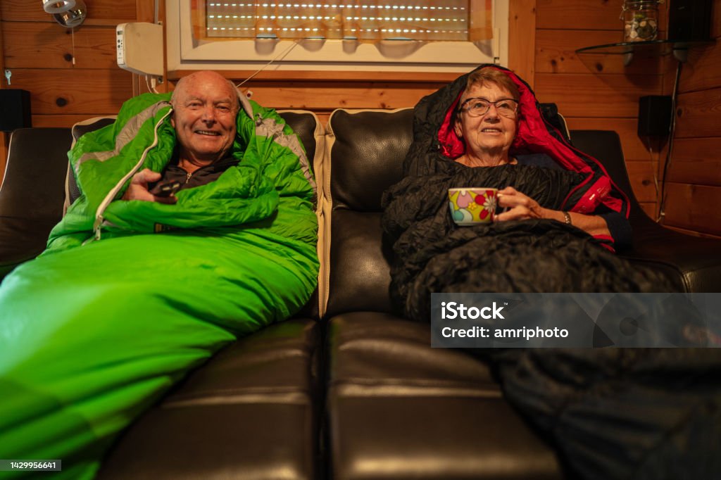Senior couple bucking the energy crisis in warm Sleeping bags at home Senior adult couple watching tv warming in sleeping bags on sofa at home in times of energy crisis, increasing electricity costs, trying to reduce heating Cold Temperature Stock Photo