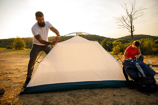 Wide shot of two friends working as a team and setting up camp for the night after a relaxing nature hike.