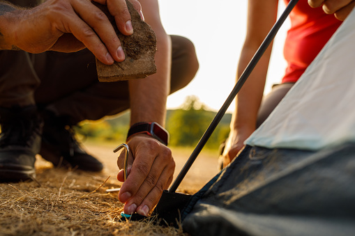 Close up shot of unrecognizable man using a rock when putting up a tent peg and setting up camp, with a friend, for the night, on the sunny meadow.