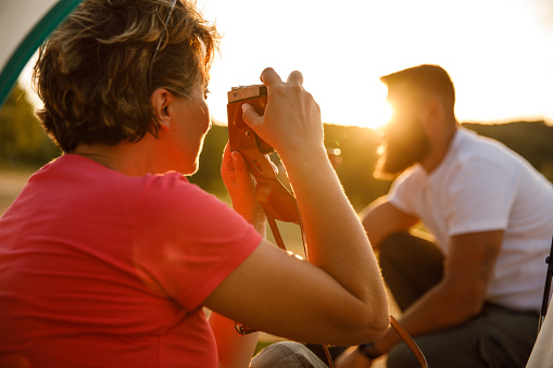 Close up shot of mature woman sitting in a tent and taking photos, with analog camera, of the beautiful golden hour sunset. Man sitting in front of the tent, enjoying picnic.